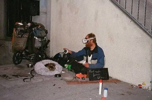 The meme is a photo of a person sitting on the street with a sign that says "Food stamps don't cover dental." The person is wearing a VR headset and has a dog sitting next to them. The image is likely meant to be humorous, implying that the person is using the VR headset to escape the harsh realities of their life on the street.